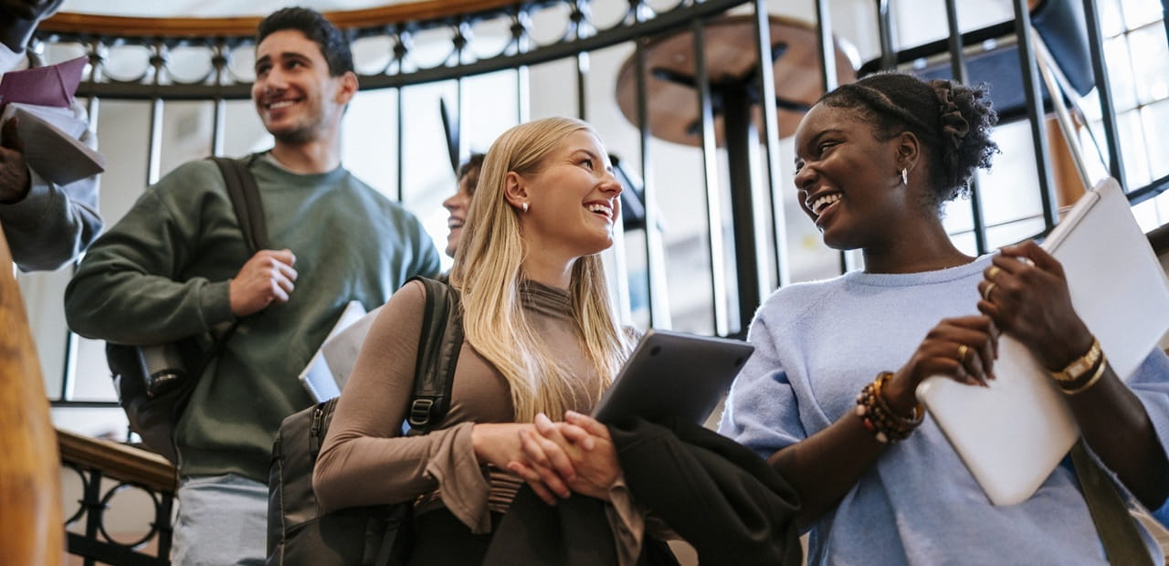 Students carrying backpacks and tablets walk through a campus building, representing the educational goals families can help support through a 529 college savings plan.