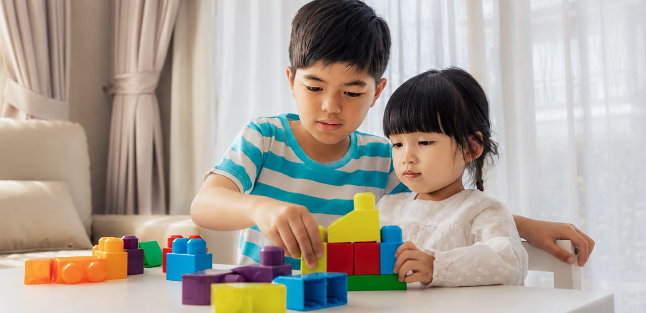 Two siblings work together to build a structure with colorful blocks at a table, illustrating how sibling relationships help form the foundation of family connections and support.