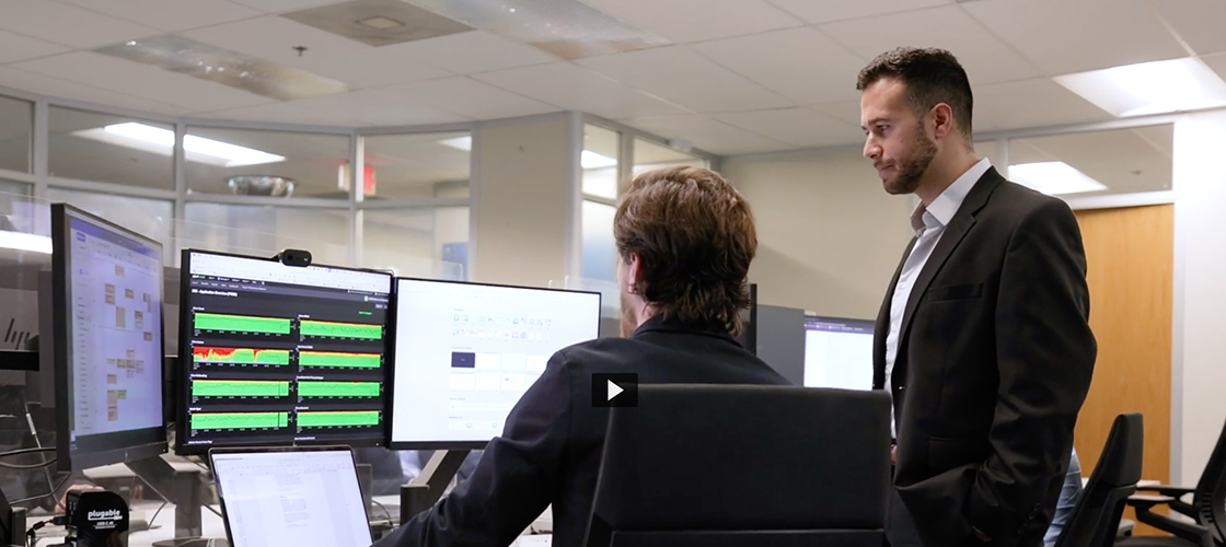 Two coworkers review application dashboards and performance metrics on multiple computer monitors in an office workspace
