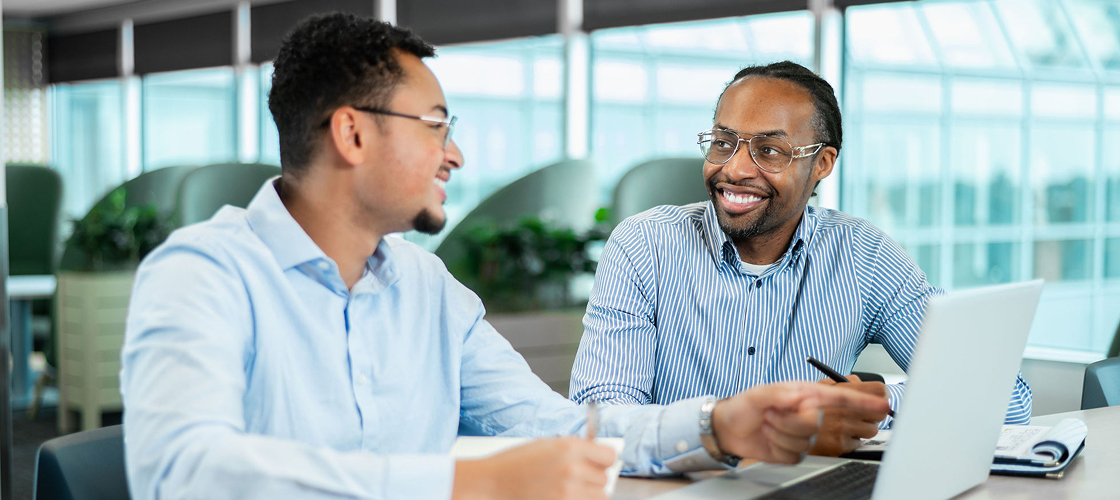 Two coworkers collaborate at a table, discussing ideas while reviewing notes and working on a laptop in a modern office.