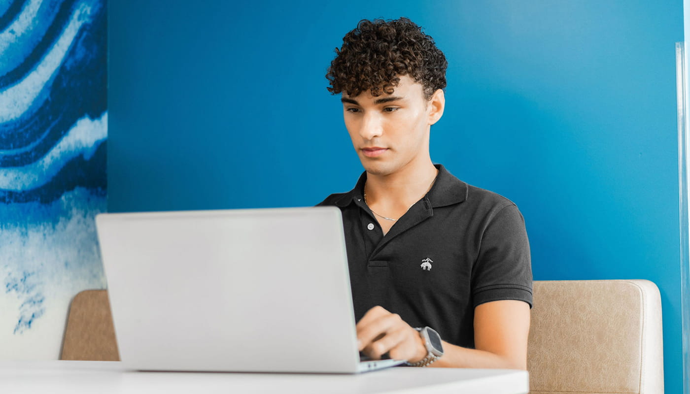 Person working on a laptop at a table in a modern office or collaborative workspace with a blue wall in the background.