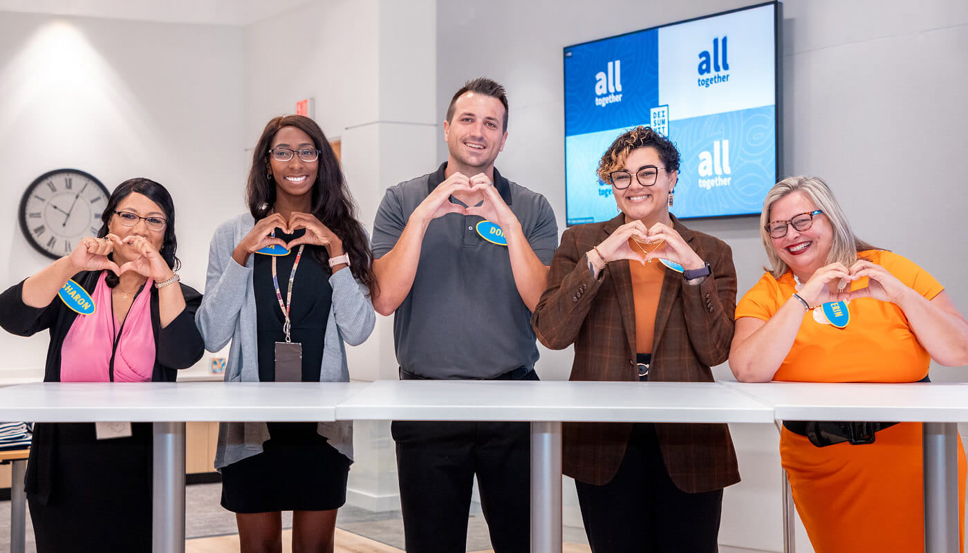 Five employees standing together in a modern office, smiling and making heart hand gestures during a company engagement or team-building event.
