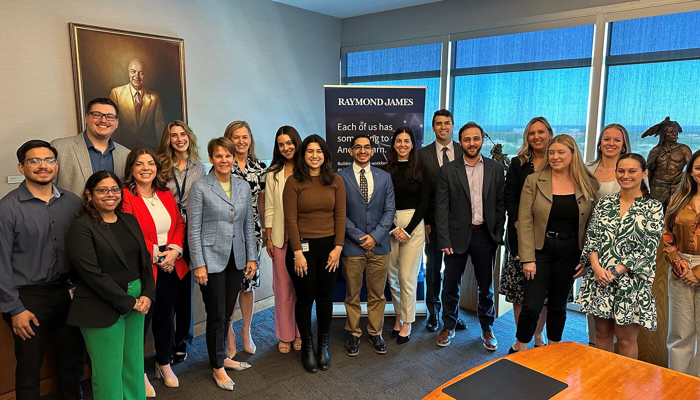 Group of employees posing together in a conference room beside a Raymond James banner during a company event, with office artwork and large windows in the background.