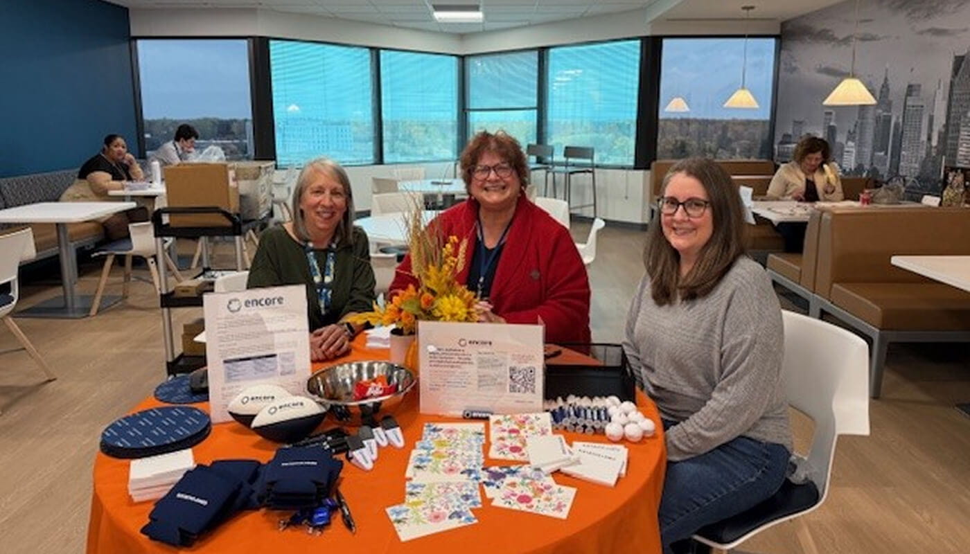 Three employees seated at an information table with Encore program materials in a company café or collaborative workspace during an employee engagement event.