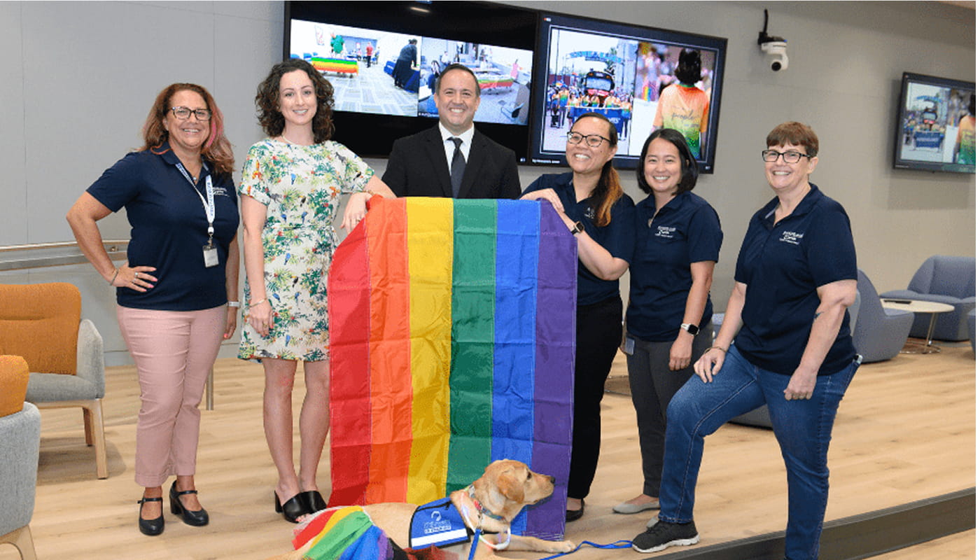 Group of employees standing together with a rainbow Pride flag and a service dog in a modern office, celebrating an employee inclusion and community event.