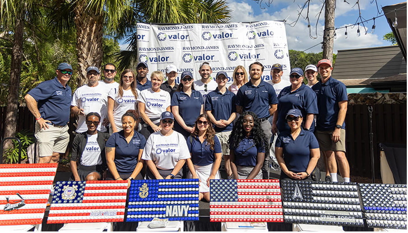 Group of employees and volunteers posing outdoors at a community service event in front of a Raymond James Valor banner, with patriotic art displays arranged in the foreground.