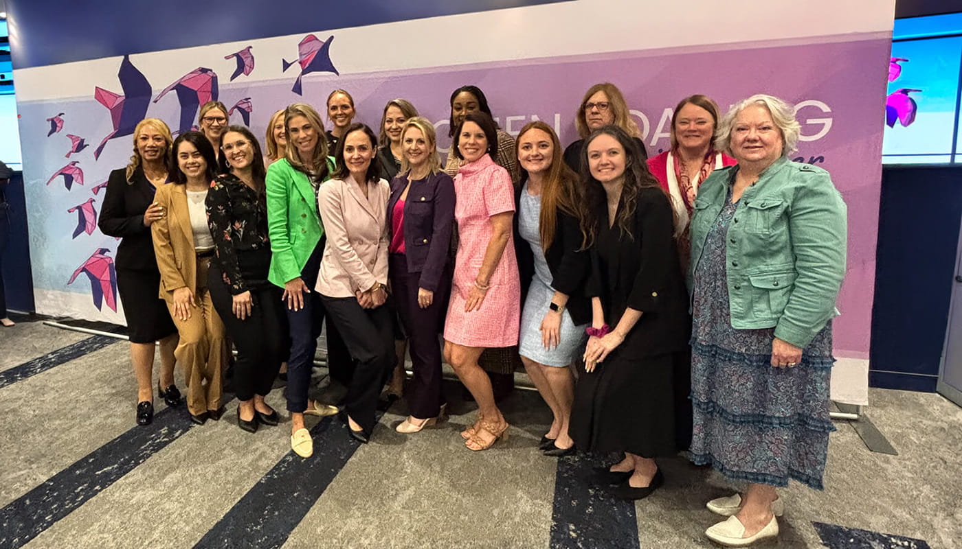 Group of approximately 16 people posing together at a professional networking event in front of a purple branded backdrop with origami bird graphics.