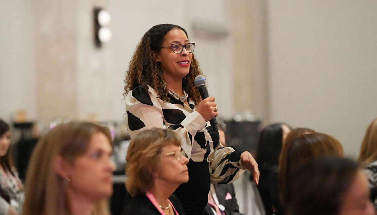 Woman speaking into a microphone while standing among attendees at a professional conference or company event, engaging the audience during a discussion.