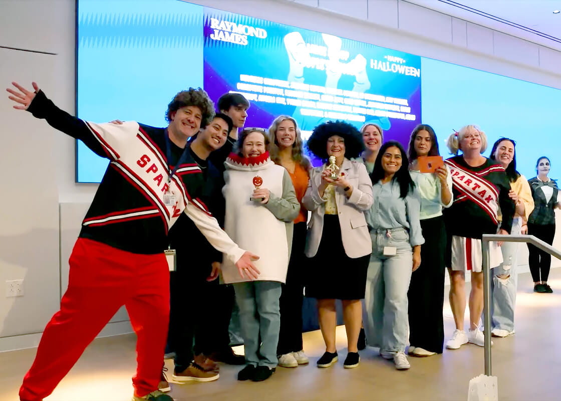 Employees wearing costumes and posing together on stage during a company Halloween celebration, smiling for a group photo in front of an event display.