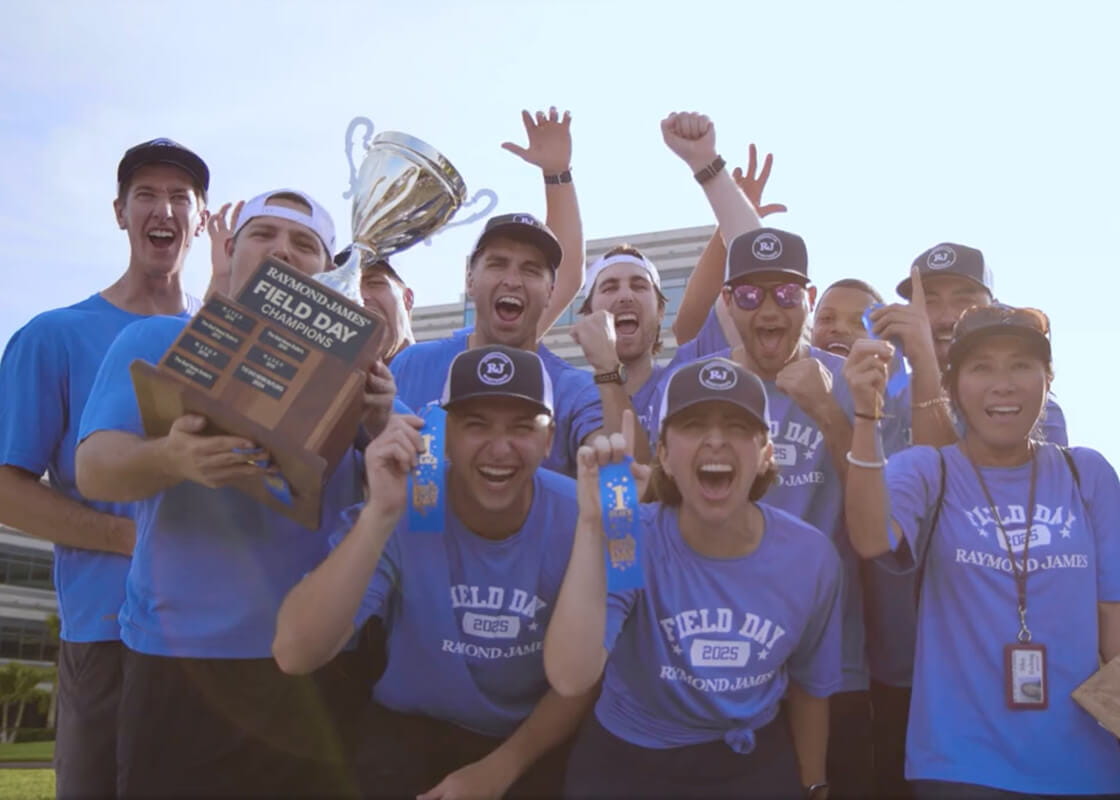 Team members celebrating outdoors after winning a company field day competition, holding a championship trophy and award ribbons while cheering together.