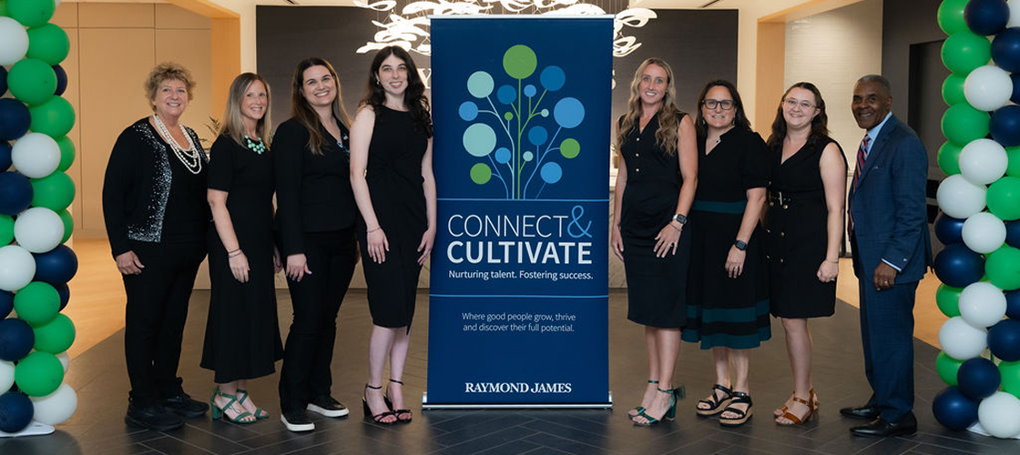 Group of employees posing beside a Connect & Cultivate program banner at a company event, framed by green, blue, and white balloon displays in a corporate lobby.