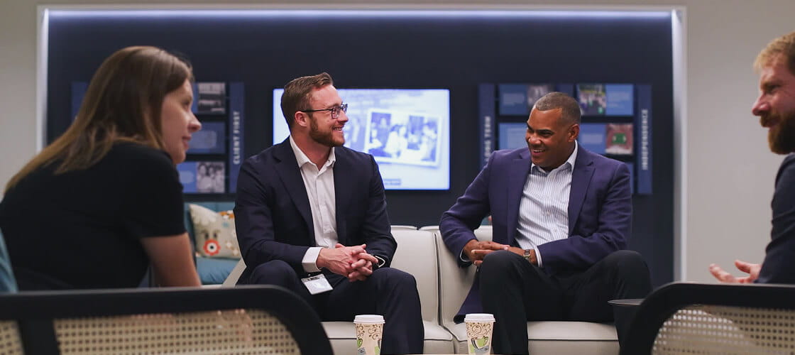 Group of professionals seated in a lounge area engaged in a collaborative discussion, smiling and conversing in a modern office setting.